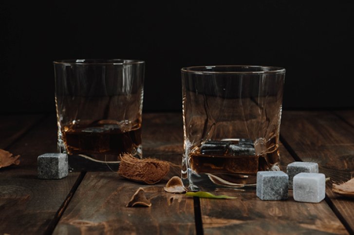 Zwei Whiskygläser mit Eiswürfeln stehen auf einem Holztisch, umgeben von Steinen und Nüssen.