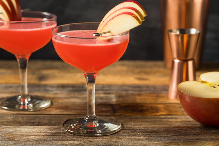 Zwei Cocktailgläser präsentieren rosa Drinks mit Apfeldeko, auf rustikalem Holztisch neben Kupfer-Shaker.