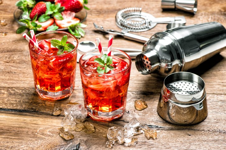 Zwei rote Cocktails mit Eis und Minze auf Holztisch, umgeben von Cocktailzubehör und Erdbeeren.
