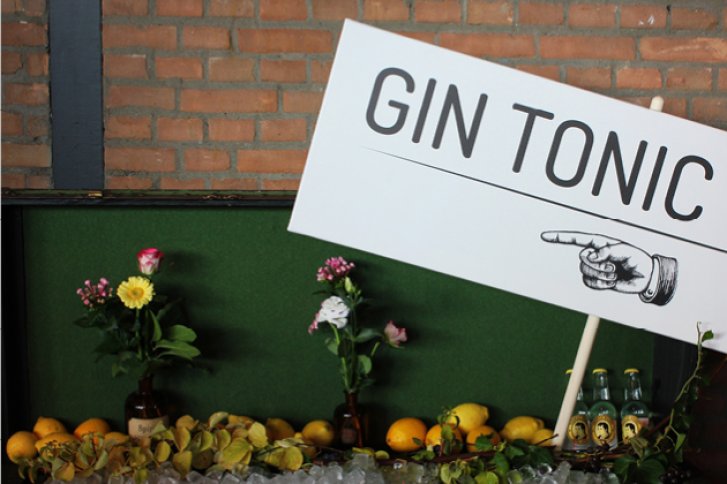 Ein Schild mit der Aufschrift „GIN TONIC“, umgeben von Blumen und Zitrusfrüchten, steht vor Ziegelwand.
