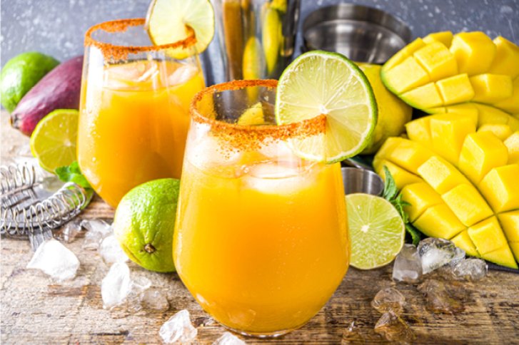 Zwei Gläser Mangosaft mit Limettenscheiben stehen auf einem Holztisch, umgeben von Limetten und Mangos.