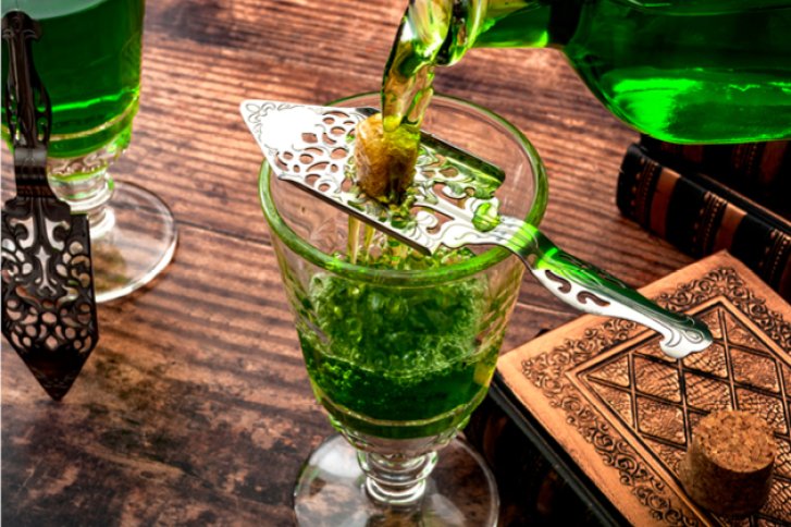 Grünes Getränk fließt in Glas mit Absinthlöffel und Zucker, auf hölzernem Tisch mit Büchern.