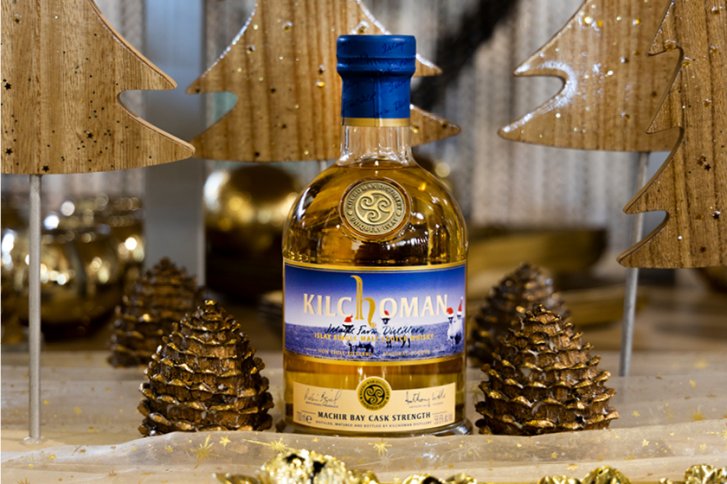 Eine Whiskyflasche steht zwischen goldenen Tannenzapfen und dekorativen Holz-Weihnachtsbäumen.