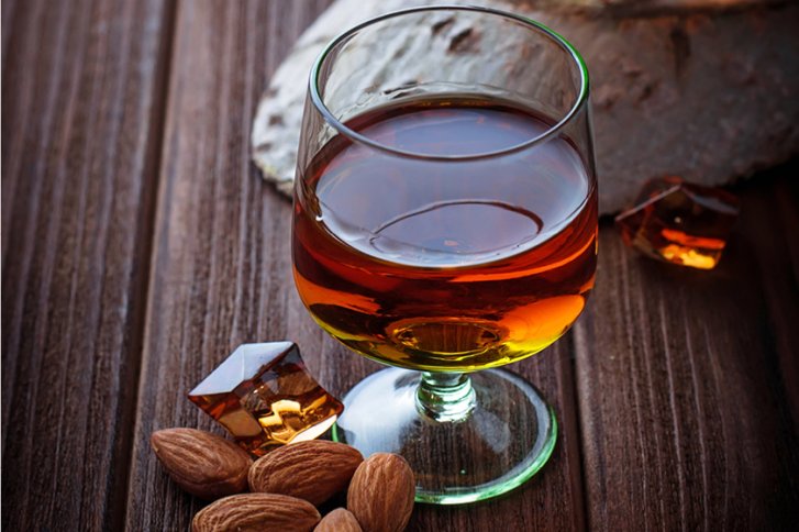 Ein Glas brauner Likör steht auf Holzoberfläche, umgeben von Mandeln und Karamellstücken.
