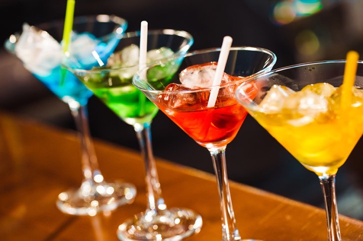 Vier bunte Cocktails stehen mit Eis und Strohhalmen auf einem Holzuntergrund in einer Bar.