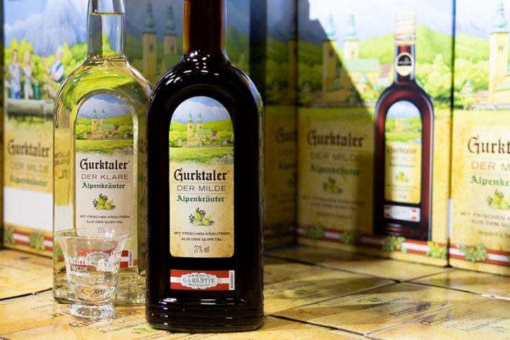 Flaschen mit Gurktaler-Alpenkräuterlikör stehen auf Kartons, umgeben von detailreichen Landschaftsverpackungen.