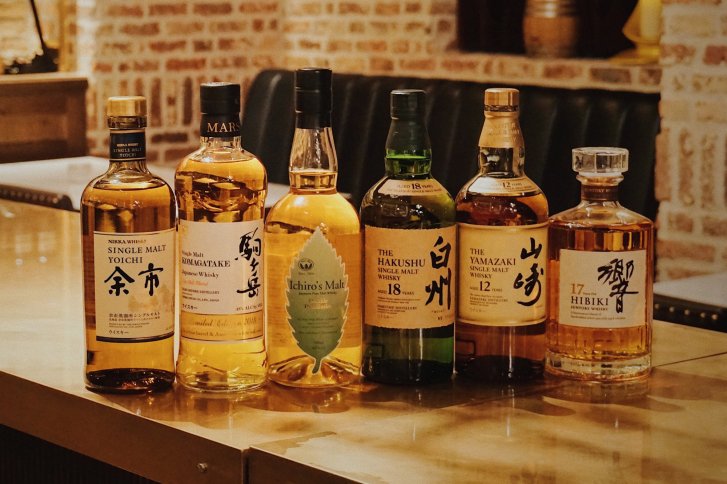 Sechs Whiskyflaschen stehen nebeneinander auf einem Holztresen vor einer Ziegelwand.