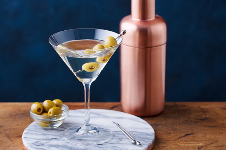 Martini-Glas mit Oliven auf Tisch, daneben Kupfershaker und Olivenschale, auf Marmortablett.