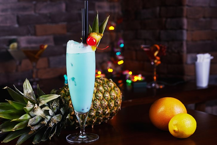 Blauer Cocktail in Glas dekoriert mit Ananas und Kirsche, vor Ananas und Orangen, bunte Lichter.