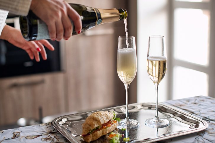Eine Hand schenkt Sekt in ein Glas neben einem belegten Brötchen auf Tablett.