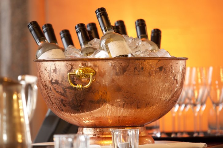Kupferschale hält gekühlte Weinflaschen auf Eis, umgeben von Gläsern im eleganten Ambiente.