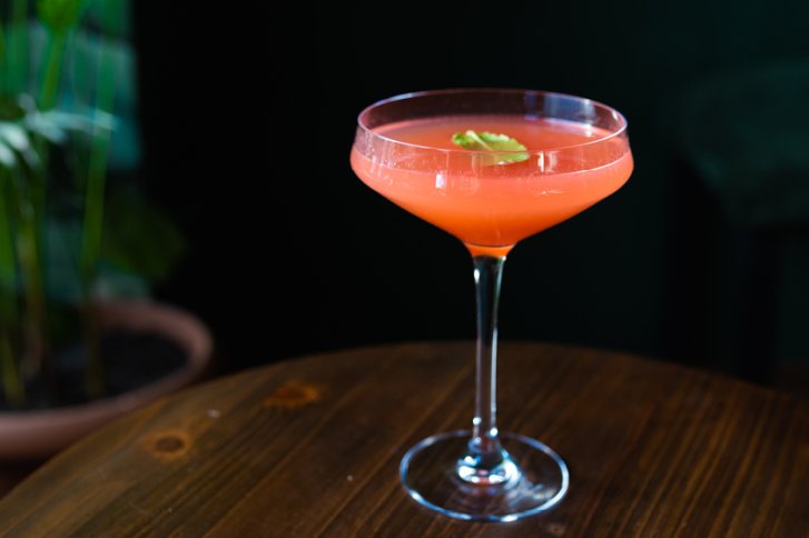 Rotes Cocktailglas auf Holztisch, dekoriert mit grünem Blatt, im dunklen, pflanzenreichen Raum.