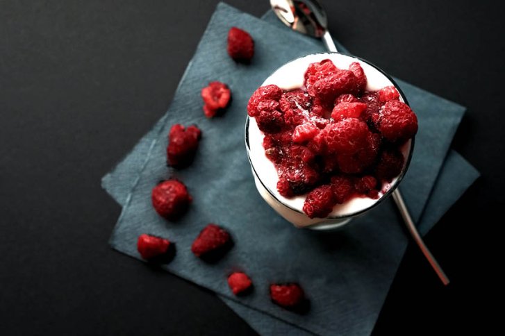 Ein Glas mit Sahne und Himbeeren steht auf dunkelblauen Servietten, daneben ein Löffel.