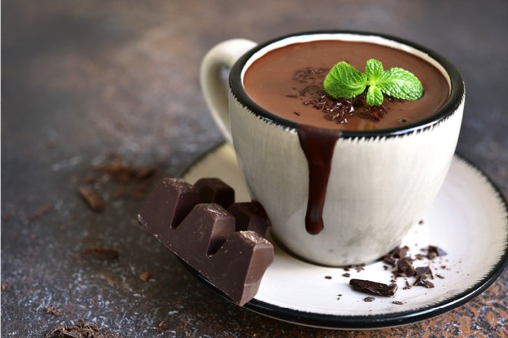 Tasse mit heißer Schokolade überläuft, dekoriert mit Minze, neben Schokostück auf Untertasse.