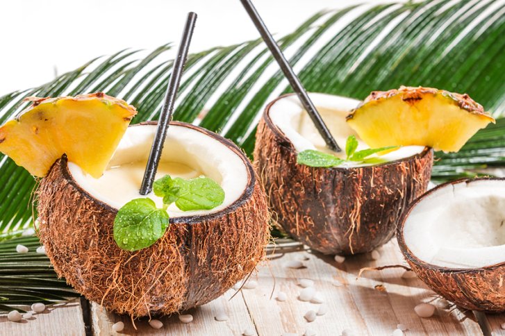 Zwei Kokosnusshälften mit Getränken, dekoriert mit Ananasscheiben, stehen auf Holz neben Palmenblatt.