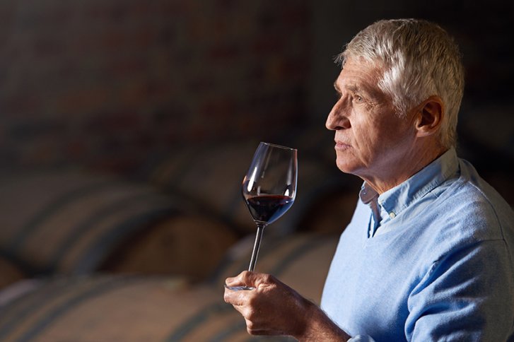 Ein Mann hält ein Weinglas, in einem Weinkeller mit Holzfässern im Hintergrund.