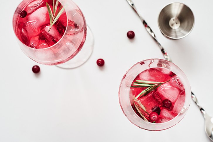 Zwei Rotwein-Gläser mit Eiswürfeln und Rosmarin, auf weißer Oberfläche, neben Cocktailzubehör.