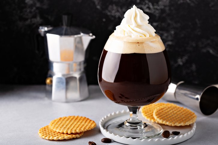 Ein Glas Kaffee mit Sahne steht auf Tisch, umgeben von Waffeln und Espressokanne.