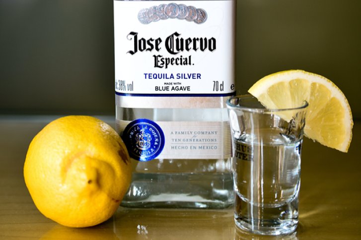 Eine Tequila-Flasche steht neben einem gefüllten Shotglas und einer Zitrone auf Holztisch.