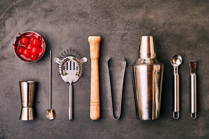 Cocktail-Utensilien liegen geordnet auf grauem Hintergrund: Shaker, Löffel, Sieb, Holzmuddler, Kirschen.