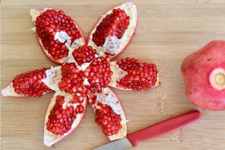 Aufgeschnittener Granatapfel entfaltet sich sternförmig auf Holzbrett, daneben ein Messer und ein ganzer Granatapfel.