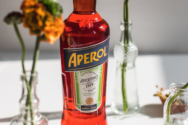 Eine Aperol-Flasche steht zwischen Blumenvasen auf einem Tisch in hellem Sonnenlicht.