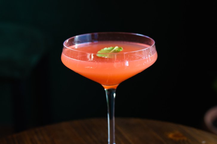 Ein roter Cocktail mit Kräutergarnitur steht auf einem Holztisch, vor dunklem Hintergrund.