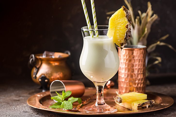 Ein Pina Colada Cocktail mit Ananasscheibe steht auf einem Tablett neben Kupferbechern.