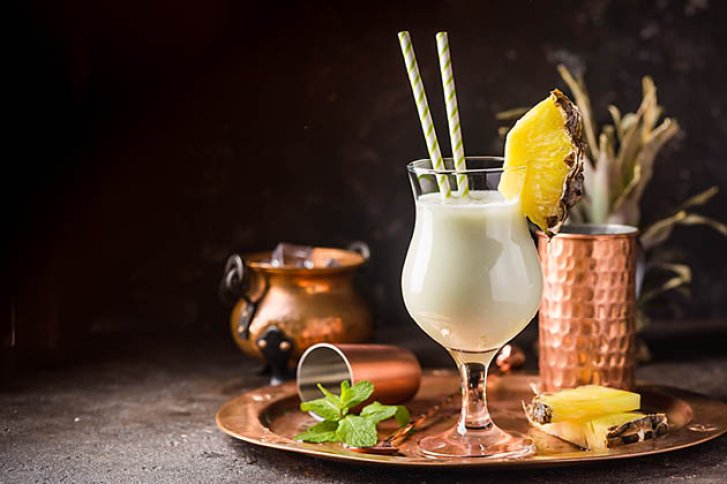 Ein Pina Colada-Glas mit Ananasscheibe steht auf einem Tisch neben Kupferbechern und Minze.