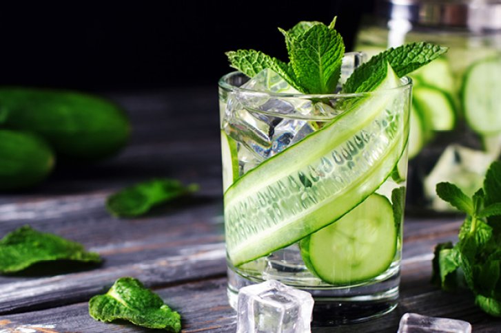 Ein Glas mit Eiswürfeln, Gurkenscheiben und Minze, auf einem Holztisch arrangiert.