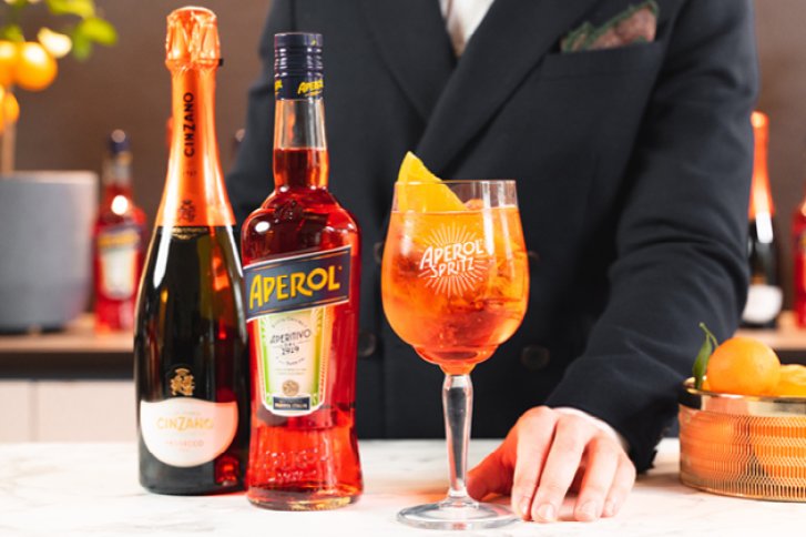 Zwei Flaschen Aperol und Cinzano stehen mit Aperol-Spritz-Glas auf Marmortisch vor Anzugträger.