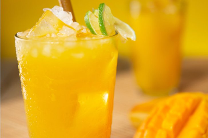 Ein gelbes Getränk mit Eis und Limette in einem Glas, umgeben von Mangostücken.
