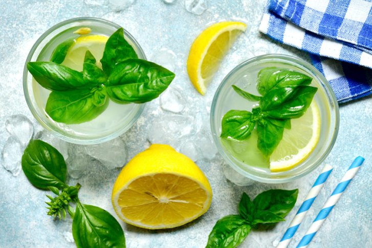 Zwei Gläser mit Zitronenscheiben und Basilikum auf Eis, umgeben von halber Zitrone, blau-weißem Geschirrtuch.