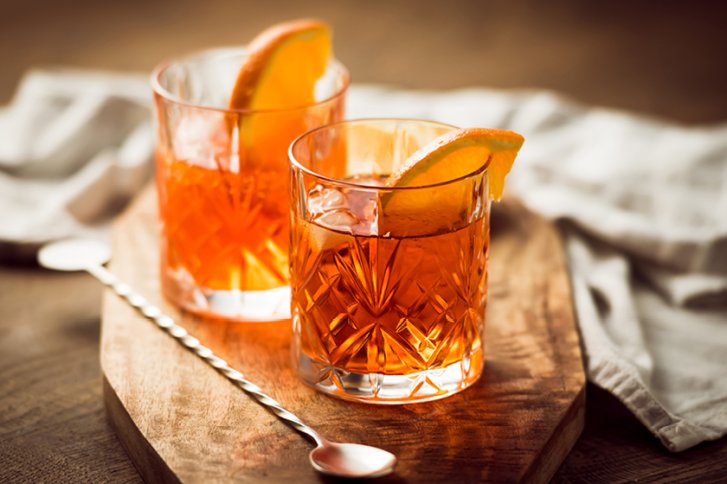 Zwei Kristallgläser mit orangefarbenem Getränk, Orangenscheiben dekoriert, auf Holzbrett, daneben Löffel.