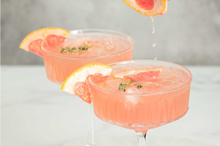 Zwei Cocktails mit rosa Inhalt, garniert mit Grapefruit und Kräutern, auf hellem Hintergrund.