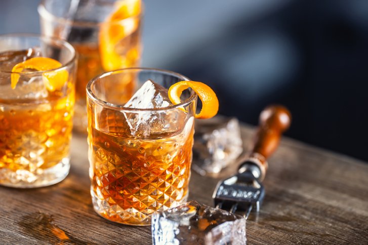 Drei Whiskeygläser mit Eis und Orangenschalen stehen auf einem hölzernen Tisch mit Eiszange.