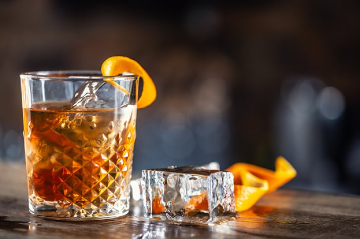 Ein Cocktailglas mit Eiswürfeln und Orangenschale steht auf einem Holztisch in einer Bar.