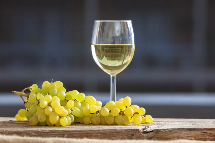 Ein Weinglas mit Weißwein steht neben grünen Trauben auf einem rustikalen Holztisch.