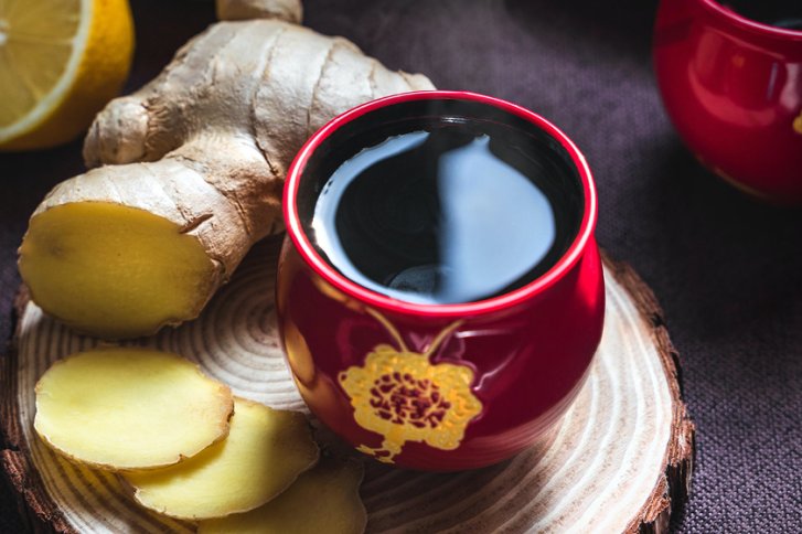 Rote Tasse steht auf Holzbrett, gefüllt mit dunklem Getränk, umgeben von Ingwer und Zitrone.