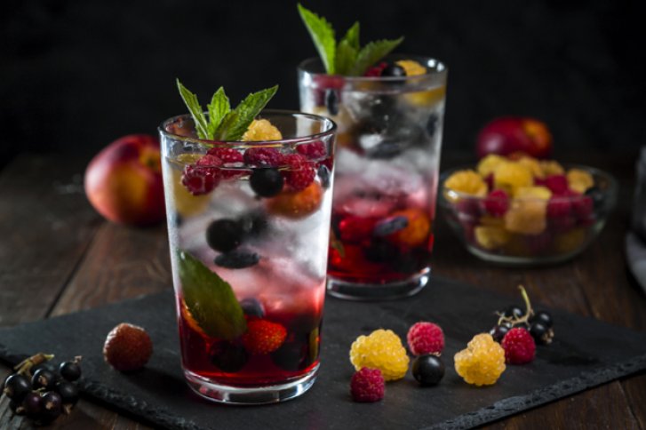 Zwei Getränke mit Beeren und Minze in Gläsern auf schwarzem Tablett, umgeben von Beeren.