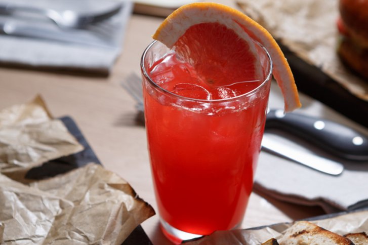 Ein rotes Cocktailglas mit Eis und Grapefruitspalte steht auf einem gedeckten Holztisch.