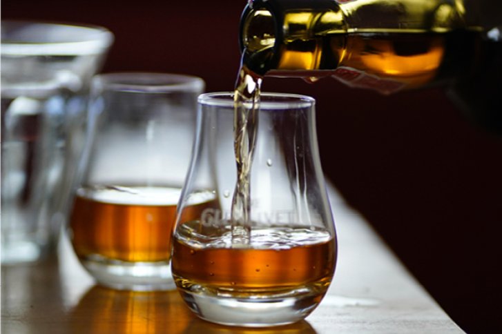 Ein Glas wird mit Whisky gefüllt, daneben stehen weitere Gläser auf einem Holztisch.