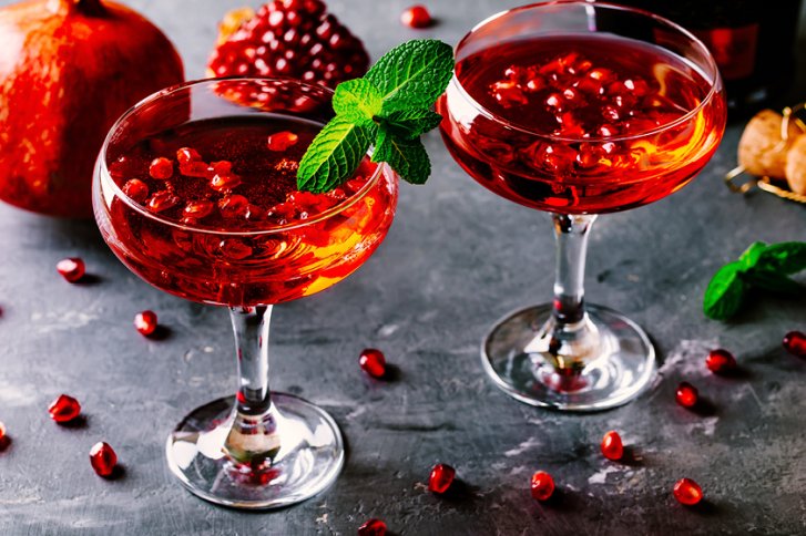 Zwei Cocktails mit Granatapfelkernen und Minze dekoriert auf grauem Untergrund, Granatapfel im Hintergrund.