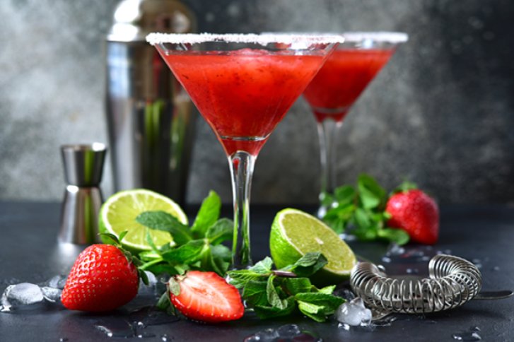 Zwei Erdbeer-Cocktails stehen auf Tisch, umgeben von Limetten, Minze, Erdbeeren, Shaker und Eis.