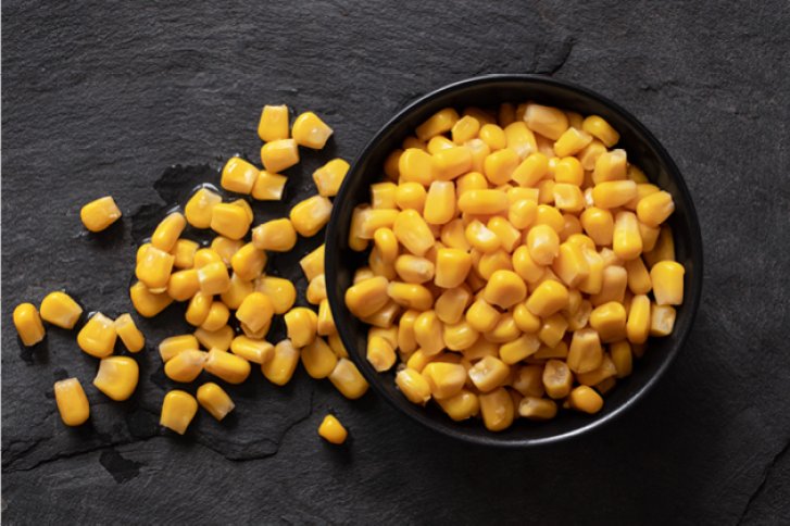 Eine Schale Mais steht auf einem dunklen, strukturierten Untergrund, einige Körner daneben.