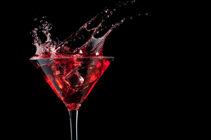 Roter Cocktail spritzt aus einem Martiniglas vor schwarzem Hintergrund, Eiswürfel sichtbar.