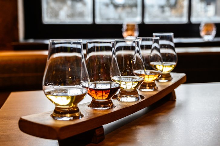 Fünf Whiskygläser stehen auf einem Holztablett, beleuchtet in einem gemütlichen, modernen Raum.