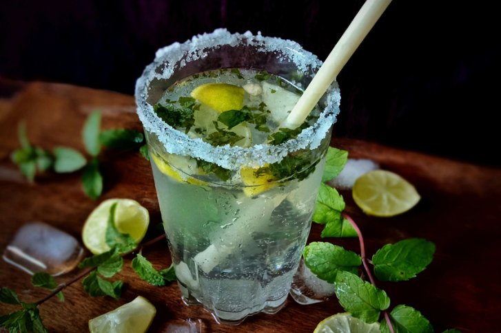 Ein Glas mit Minzblättern, Limetten und Zuckerrand steht auf einem Holztisch.