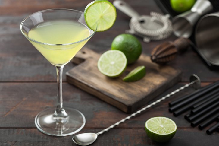 Cocktailglas mit gelbem Getränk, Limettenscheibe am Rand, auf Holztisch mit Barutensilien.