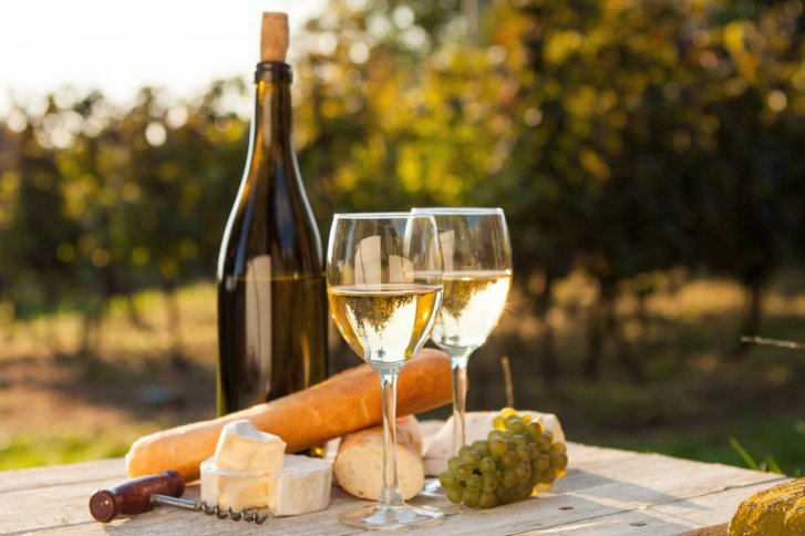 Zwei Weingläser und Flasche stehen auf Tisch mit Baguette, Käse, Trauben im Freien.